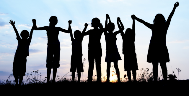 A photograph showing a silhouette of some children with a sunset behind. The children are holding hands.