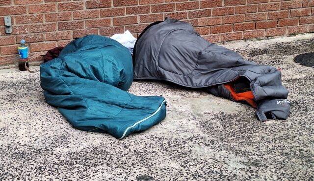 A photograph showing two people sleeping in sleeping bags on a tarmac surface.