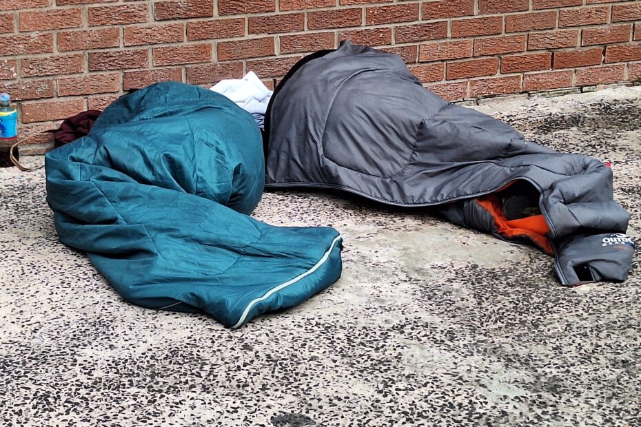 A photograph showing two people sleeping in sleeping bags on a tarmac surface.